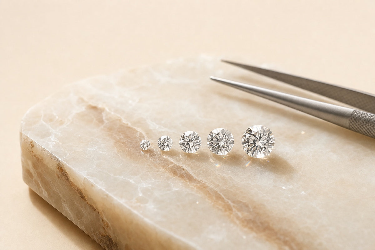 Cinq diamants de synthèse ronds brillants de tailles croissantes alignés sur une dalle de marbre crème, à côté d'une pince de gemmologue
